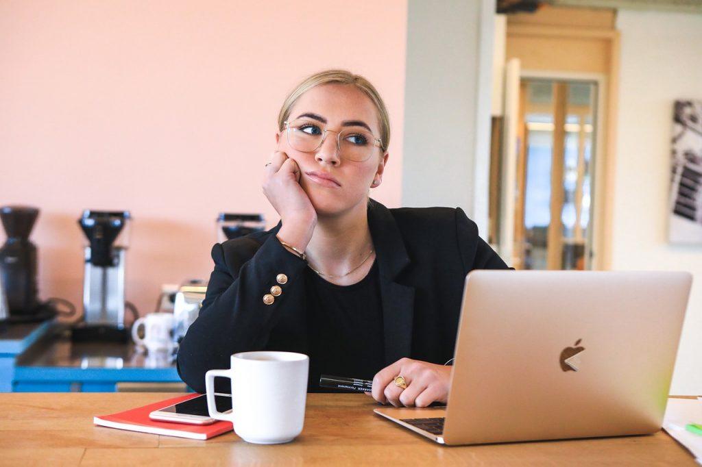 A person with glasses sits at a table in front of an open laptop, resting their chin on their hand. They appear thoughtful, perhaps contemplating strategies to overcome procrastination. A coffee mug, smartphone, and notebook are also on the table. The background shows a kitchen area.